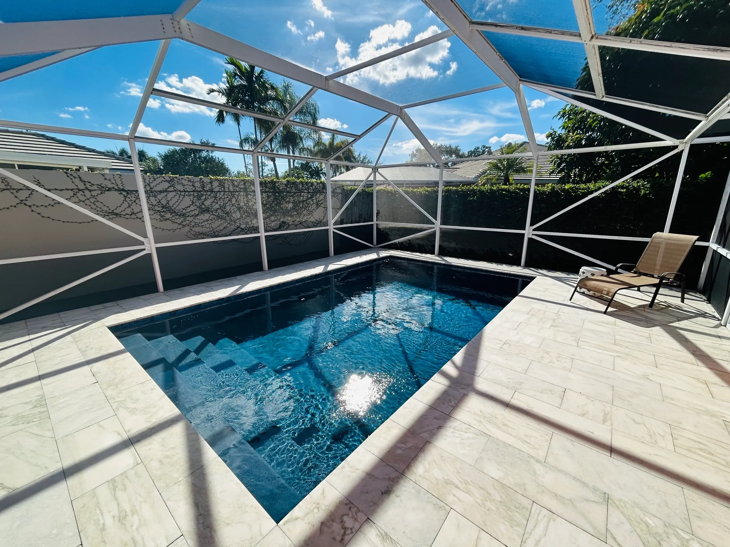 Modern pool area with marble pavers, enclosed by a white screen, featuring a sunlit pool, steps leading into the water, and a lounge chair beside the pool.