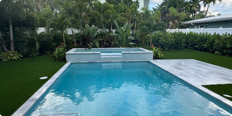 Luxurious swimming pool with spa, surrounded by lush tropical landscaping and artificial turf, highlighting custom hardscaping for outdoor spaces.