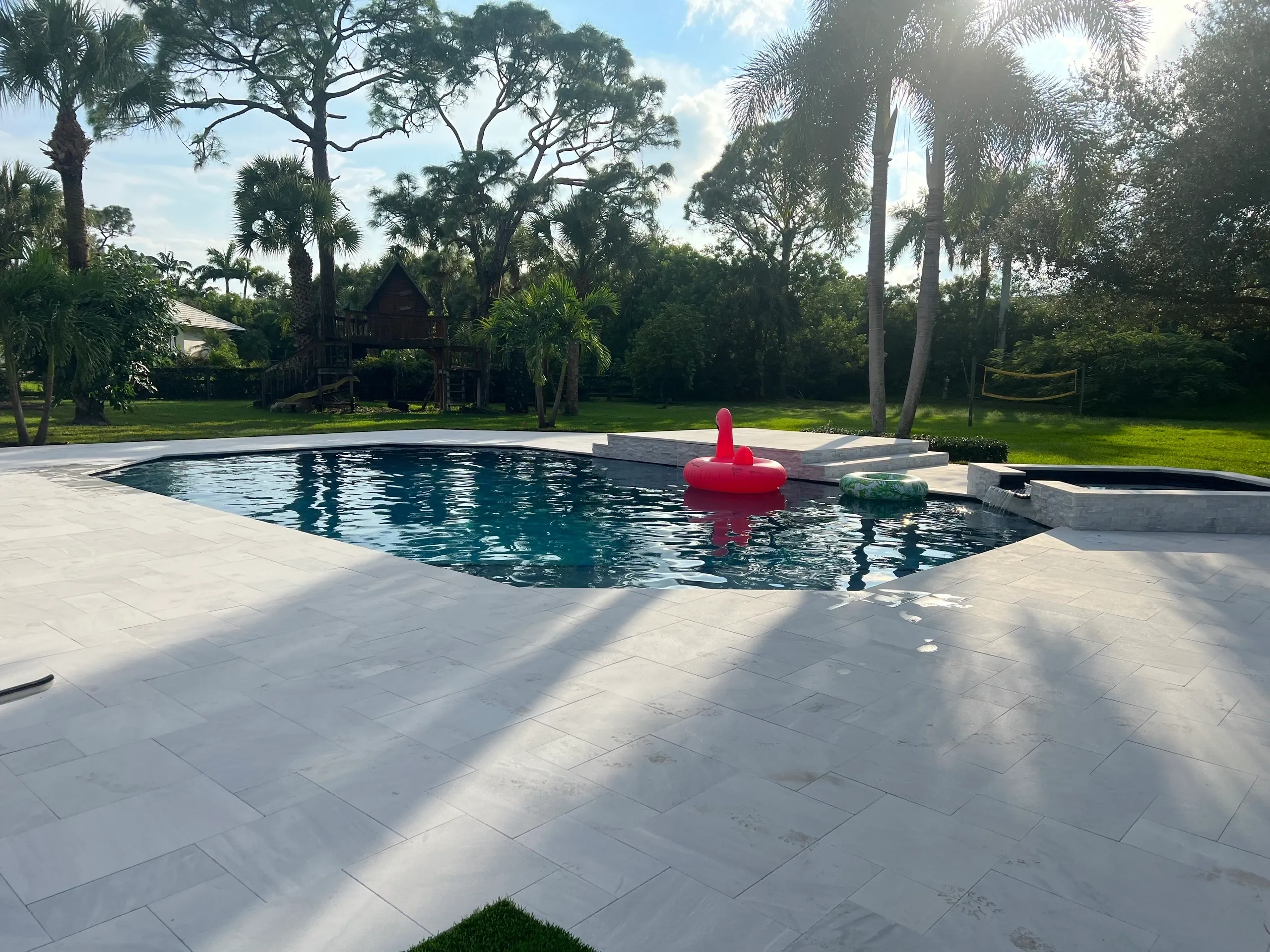 Swimming pool surrounded by elegant white pavers, featuring a pink inflatable float and a green inner tube, set in a lush backyard with palm trees and a wooden play structure in the background.
